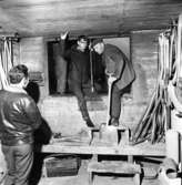 HSB´s verkstadslokaler. Fyra män ses på bilden. De tar sig in i källarrummet genom att klättra genom ett hål i väggen. I rummet finns en verktygslåda, grepar, räfsor och piasavakvastar. Den yngste mannen håller i en räfsa och den äldsta håller i en spade.
(HSB betyder Hyresgästernas sparkasse- och byggnadsförening)