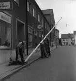 Här ordnas det inför konungens ankomst. Fyra män ska sätta upp en flaggstång.
Kung Gustaf Vl Adolf ska komma på Eriksgata 12 september.
Järntorgsgatan. Butikerna skyltar med armatur, glasögon och guld.