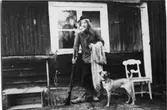 Jägare utanför sin stuga. Hans hund står bredvid honom.
Mannen håller ett gevär i ena handen och har möjligen ett fisknät över andra armen.
En trälåda, en grep och en stol står efter husväggen.