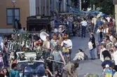 Arbogakarnevalen. Paraden har nått Rådhusgatan. Bilar, dansare och orkestrar. Mitt i bild ses Arboga Blåsorkester iklädda sombreros. Bostadsbolaget Lunds deltar med sin gamla bil. Publiken kantar gatan.