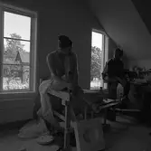 Keramikerparet Kerstin Hörnlund och Gösta Gräs i ateljén i en ågård. Kerstin sitter på en arbetsbänk. Nedanför henne står en gjutform. Gösta sitter på en hög pall. Bakom honom syns keramikföremål.
Läs Kerstins text 