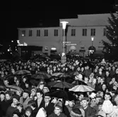 Publiken vid Lucia-kröningen på Stora torget. Många har paraply. Scenen står framför rådhuset. Skyltarna för Sture-Bio, Nerikes Allehanda och Frisör syns i bakgrunden.