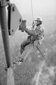 Ossian Möller på jobbet. Arbete på hög höjd.
Man i mast. Han sitter på en sits som hänger i en wire. Han bär arbetskläder och skyddshjälm. Långt under honom ses skogen, en väg och några byggnader.