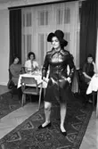 TVB ordnar modevisning och bjuder på kaffe och landgång. Ylva Martinsson visar hatt och kappa. Vid borden sitter, från vänster: Ingrid Östling, Anne-Marie Andersson och Eva Heiner.
Eftersom Sture Melander ses på en bild i sammanhanget, kan man gissa att kläderna kommer från Öhrman & Melander.