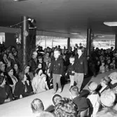 Modevisning hos Arboga Bil AB. Här kommer tre barn i dubbelknäppta kavajer och långbyxor.
Gott om publik i bilhallen. Människor har ytterkläder på sig. Lokalen är pyntad med björklöv och ballonger. Det kan vara bilfirmans 1-årsjubileum.