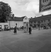 Nytorget.
Människor sitter på bänkar vid fontänen. En telefonkiosk är placerad mitt på torget. Två flickor äter glass. Butiker på Nygatan ses, bland andra Tobaksaffären. I förgrunden hänger en GB-glassflagga.