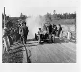 Reproduktion Röfors. En man kör en bil eller en traktor. Det kommer ett stort rökmoln bakom fordonet. Människor står i vägkanten och tittar på. Bilden är tagen på landet. Byggnader skymtar i bakgrunden.
