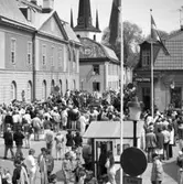 Riksdagsjubileet. I Arboga firas 550-årsminnet av Sveriges första riksdag. Många höga politiker samt medlemmar ur kungafamiljen gästar Arboga denna dag. Motorcykelburna poliser, en polisbil och patrullerande poliser sköter säkerheten och håller kontroll på den stora publiken. Många människor har samlats på Rådhusgatan. Vid dörren till Arboga Tidning ses ett par män i Arboga Blåsorkester med sina instrument. Någon tittar ut genom ett öppet fönster i Musicum. I bakgrunden ses tornet på Heliga Trefaldighetskyrkan.