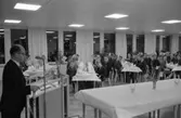 En talare på Stureskolans invigningsfest. Vid dukade bord sitter festklädda människor. Några unga pojkar sitter vid de närmaste borden.
Påskliljor i vas.