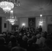En sångkör uppträder i kommunfullmäktigesalen på Rådhuset. Kören består av både män och kvinnor. En stor publik lyssnar under kristallkronorna. På väggen hänger en stor målning av kung Gustav ll Adolf.
sjunga