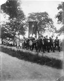 Söndagsskolan har fest. Barn och vuxna tågar på en grusväg nära vattnet. Främst går en blåsorkester. En man bär en fana.
Fotografiet är gjort som ett vykort med adressrader på baksidan.
Baksidan på fotografiet ses AKF 12072
