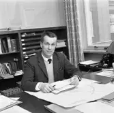 Tage Schyberg, byggnadschef.
Han sitter vid sitt skrivbord. Där ligger en ritning och en hög med post. En telefon står nedanför fönstret. Bakom ryggen har Tage en bokhylla.