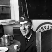 Arne Fallkvist/Arne Fallqvist har kafferast på taxistationen. En kaffepanna och en kopp står på bordet. Arne bär båtmössa. Han har ett plåster under ögat. Troligen skadade han sig under en ishockeymatch. Arne är en mycket duktig målgörare.
Porträtt av man i arbetsuniform.