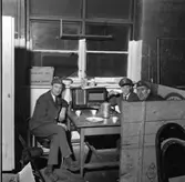 Kafferast på taxistationen. Chaufförerna sitter vid bordet. Till vänster Stig Johansson. Han bär ett sorgband på kavajslaget. Mitt emot honom sitter Karl-Erik Larsson i skärmmössa. Bredvid honom sitter Arne Fallkvist/Fallqvist. Arne har ett plåster under ena ögat. Troligen har han skadat sig när han spelat ishockey.
De har en kaffepanna och koppar på bordet. Där finns även en stor radioapparat. På fönsterkarmen står en pappkartong med texten 