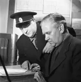 Taxistationen. Två män på kontoret. Chaufför Halvar Hellkvist i skärmmössan med taxi-märket. Den andre mannen är Hilmer Jansson alternativt Hilmer Jonsson. (Han har smeknamnet Bergvall, efter någon idrottsman)