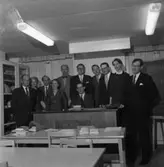 TBV, högskolekurs.
Män och kvinnor är samlade runt katedern i ett klassrum. På bänkarna ligger uppslagna böcker. En svart tavla syns bakom gruppen.
Det är lågt i tak i rummet, vilket ger känslan av att de befinner sig i en källarlokal. Lysrörsarmatur.
Tjänstemännens BildningsVerksamhet