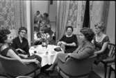 Televerkets personal. Uppklädda kvinnor sitter vid ett runt bord.  På bordet står kaffekoppar, glas och en blomkruka med en Amaryllis i. Där ligger även ett glasögonfodral, en tändsticksask och en aftonväska.
Kvinnorna är, från vänster: Greta Östling, Britta Conradsson/Konradsson, Alice Ståhl (med ryggen mot spetsgardinen), Kerstin Pettersson (med tvåradiga halsbandet) och okänd kvinna (som även syns i den stora spegeln mellan fönstren).