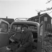 Tore Andersson i Mossen.
Han sitter i bakluckan på sin Amazon kombi och har en krimmelmössa på sig. Bakom bilen ses två boningshus.