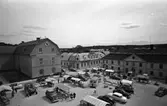 Torghandel med flera torgstånd och bilar på Stora torget. Människor promenerar mellan stånden.
I bakgrunden ses Rådhuset och innegården, till vänster. Möjligen 