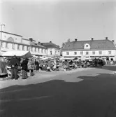 Torghandel, mest blommor i stånden. Män i hattar och kvinnor i dräkter. Platsen är Stora torget. I bakgrunden ses 
