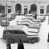 Det är mycket snö och svårt att parkera på Stora torget. Bilarna står tätt. Människorna är rejält påklädda. I bakgrunden ses Lundborgska fastigheten med café Saga