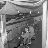 Kvinnorna arbetar vidare i telefonväxeln trots branden på Televerket på Nygatan. En presenning är uppspänd över växelbordet och personalen. Nakna glödlampor hänger från taket.
Läs om Telefonen i Arboga och branden på Televerket 1956 i Arboga Minnes årsbok 1993.