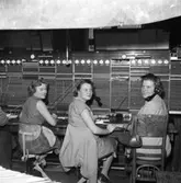 Televerket på Nygatan, efter branden. En presenning hänger i taket för att skydda växeln från det vatten som användes i släckningsarbetet. Växeltelefonisterna arbetar med en telefonlur hängande vid vänster öra. Klockorna, på rad, visar hur många perioder samtalen pågått.
Från vänster: Britt Johansson, okänd och Mary Pettersson.
Läs om Telefonen i Arboga och branden på Televerket 1956 i Arboga Minnes årsbok 1993.