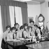 Ungdomens Röda Kors.
Bakre raden: Monica Karlsson, Elisabeth Högfeldt, okänd, Gerd Elöw
Sittande: Eva Parli (mötessekreterare), Inga Johansson med ordförandeklubban, Margareta Thegström och okänd.
Röda Korset
Unga kvinnor i skjorta, slips och basker. De har möte. I lokalen finns ett porträtt på kung Gustav V och en moraklocka. På bordet finns ljusstakar, en flagga och en vas med tulpaner