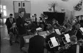 Ungdomsorkestern/Blåsorkestern uppträder. Dirigenten och de flesta av musikerna är klädda i uniform. Notställen är uppställda. I bakgrunden står ett piano.