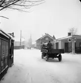 Östra Hamngatan i snöfall. En man kör med häst och kärra förbi Texacos bensinstation. På höger sida ses hamnmagasin. I bakgrunden kan man skönja Heliga Trefaldighetskyrkan.