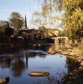 Vy mot Kapellbron och Heliga Trefaldighetskyrkan. Lågt vatten i Arbogaån. Kvällssol på sommaren.
Bilden är tagen från en brandgata från Storgatan (södra sidan om ån).