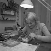 Ann-Britt Gustafsson, årets Lucia.
Ung kvinna som sitter vid skrivbordet. Framför henne ligger en isärtagen teknisk apparat. Hon håller en skruvmejsel i ena handen. Fler mejslar liggar på skrivbordet. En skrivbordslampa syns. På väggen hänger en bokhylla, modell String, med böcker, prydnadssaker och en krukväxt.