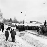 Invigning av ombygda vägen till Örebro. Trots vinter och snö har många människor samlats för att vara med när bandet klipps. Någon har kameran med sig. Bakom publiken, står flera fordon och väntar på att få köra; först två personbilar och sedan flera bussar. I motsatt riktning är en lastbil parkerad. På taket till förarhytten står en man. Han håller en lapp, med sitt tal, i handen. En stege är rest mot förarhytten.