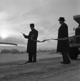 Bandet klipps! Örebrovägen öppnas efter ombyggnad. En man med vinterrock och krimmelmössa håller i saxen. Bredvid honom står en man i vinterrock och hatt. Bakom honom syns motorhuven på en lastbil.