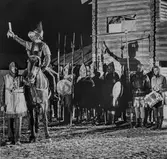 En scen ur skådespelet Giv oss fred. En riddare, i rustning, till häst. Män med sköldar och hillebarder. Bredvid hästen står kusken Gustav Jansson.
