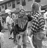 Höstmarknad på Järntorget. Två unga män samtalar. Den vänstra är Robert Andersson. Fler ungdomar i bakgrunden, längst till vänster står Peter Berg. Ett marknadsstånd med hemkokta karameller.
