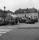 Torghandel. Höstmarknad på Stora torget. Frukt och grönsaker säljs. En familj handlar. Ett barn sitter i barnvagn. Människor i rörelse. Det är blomsterplanteringar på torget. Bilar parkerade till vänster. I bakgrunden ligger Duka-butiken.