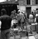Höstmarknad på Järntorget. Människor besöker torgståndet med korgar. En ung kvinna har en korg på armen och en plånbok i handen.
Byggnaden i bakgrunden är 