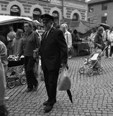 Höstmarknad på Järntorget. Bland människor och marknadsstånd går en man iklädd Frälsningsarméns uniform. Han har en insamlingsbössa i handen.
Byggnaden i bakgrunden är 