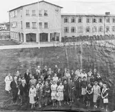 Lärare framför Gäddgårdsskolan.
Längst till vänster, i ljus kappa, står Karin Stenström (folkskollärarinna). Intill henne står Nils Karlsson (slöjdlärare). Mannen bredvid honom är Arne Bielke (musik-och bildlärare).
Närmast framför dessa står, från vänster: Folke Pettersson (folkskollärare), Alice Hedlund (småskollärarinna, i spräcklig blus), okänd kvinna i glasögon och Karl-Olov Karlsson (folkskollärare).
Framför dessa fyra står två män; Bror Gustavsson (rektor) och Gösta Kjellmert (folkskollärare). Kvinnan framför Gösta är Karin Booberg (talpedagog).
I bakre raderna står två herrar med fluga. Den med randig fluga heter Albert Svensk (folkskollärare). Bakom honom, i mörk fluga, står Lennart Karlsson (folkskollärare). Intill Lennart står Walter Falk (folkskollärare) och framför Walter står Erik Karlsson (slöjdlärare).
Framför Albert, med randiga flugan, står Arvid Imark (folkskollärare). Kvinnan bredvid honom heter Lena Nohlgren (folkskollärarinna). Framför Arvid står Inga-Britt Nannfeldt (folkskollärarinna). Framför henne, i dräkt, står Barbro Ståhl (småskollärarinna). Intill Barbro, i mönstrad klänning, står Naemi Ageborg (folkskollärarinna) och bredvid henne, i ljus klänning och handväska, står Marianne Holmberg (folkskollärarinna). Mellan Naemi och Marianne syns Borgil Márcör (småskollärarinna). Bakom henne, i randig blus, står en oidentifierad lärarinna. Bakom henne, i mörk jacka, står Eva Möller (småskollärarinna). Mannen bakom Eva är Gottfrid Lönnberg (folkskollärare).
Den högra av de två kvinnorna med ljusa handväskor, på mitten av främre raden, heter Britta Jönsson (gift Sundkvist, småskollärarinna). Bakom henne står Barbro Karlsson (småskollärarinna). Intill Britta står Birgitta Lundman (gift Göransson, småskollärarinna). Bakom Birgitta, i spräcklig kavaj, står Karl Porath (gymnastiklärare). Bakom honom, i ljus kappa, står  en okänd lärarinna. Kvinnan till vänster, bakom henne, är Karin Eriksson (småskollärarinna).
Intill den oidentifierade lärarinnan, i ljus kappa, i bakre raden, står sex lärarinnor i rad. De är från vänster: Greta Markusson, i mörk kappa och vit krage (folkskollärarinna), oidentifierad lärarinna i ljus kappa, kvinnan i hatt är Dagmar Rydbeck (småskollärarinna), bredvid henne står Karin Björk (textilslöjdslärarinna), bredvid henne, i kofta och kjol, står Eivor Pettersson (småskollärarinna) och längst till höger Anna Arvidsson (småskollärarinna).
I raden framför dem, står tre kvinnor. Den första står snett framför kvinnan i hatt, hon har en spräcklig blus och glasögon och är oidentifierad. I samma rad, fast långt åt höger står två kvinnor i mönstrade klänningar. Hon med båtringningen heter Gun Martinsson (småskollärarinna) och hon med vita kragen är Nanna Sander (folkskollärarinna).
I den främre raden står sju kvinnor. Vi börjar med kvinnan i spräcklig dräkt och pärlhalsband, Karin Gustavsson (gift med rektor Bror Gustavsson, folkskollärarinna). Intill henne, i mörk dräkt, står Aina Ungvall (småskollärarinna). Bredvid henne, i mönstrad blus och ljus kjol, står Britt-Inger Axelsson (småskollärarinna). Framför henne, i svart klänning med medaljonghalsband, står en oidentifierad kvinna. Bredvid henne, i ljus dräkt, står Gerda Svensson (småskollärarinna). Bakom Gerda syns en oidentifierad kvinna. Längst till höger står Wivan Söderberg (småskollärarinna).
Gruppfoto.
Avfotograferad påsiktsbild.