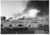 Det brinner i Elektriska A-B Skandia! Lågorna slår högt över taket på byggnaden. Tre brandmän står på taket.