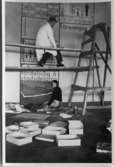 Två personer bygger en utställning på Arbogautställningen 1935. En man iklädd skyddsrock sitter på en enkel byggställning. På golvet sitter en kvinna. Båda arbetar på samma vägg. Kartonger ligger utspridda på golvet.