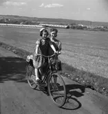 En kvinna leder en damcykel som en liten flicka åker på. Bilden är tagen på dåvarande Nässjövägen i backen upp mot Ekhagen och i bakgrunden syns A6 förrådsbyggnader, Vättern och Jönköping.