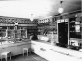 Handelsbod, diskar in vinkel. Kassaapparat i hörnet, en våg på varje disk. Specerier på hyllorna till vänster och tygrullar till höger.
Lanthandel