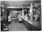 Handelsbod med sju personer i personalen. Alla är klädda i arbetsrock. Här säljs tvättmedlet Tvätt-Björn, julgransljus och även bananer. Varorna är staplade i pyramider på disken.
Lanthandel
(Den första bananlasten kom till Sverige i november 1944)