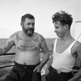 Två män ombord på ett fartyg. En äldre man med tatueringar samt en yngre man.