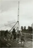 Sättning av första kontaktledningsstolpen på Trafikaktiebolaget Grängesberg - Oxelösunds Järnvägar, TGOJ:s spår. Arbetslaget tar en paus medan ett ångtåg passerar den 12 juni 1952.