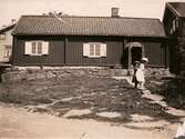 Porträtt av två flickor framför det troligen äldsta huset i Fässbergs by. Fässberg Västergård 6, Fässberg 1:34. Fotografiet är taget ca 1910. Flickan i den stora hatten är fotografiets ägares farmor Edit Olsson, född 1903 (gift 1923 Andersson).