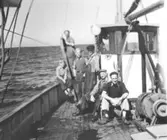 Bild 1:Stående längst bak Erik Mårtensson, därefter två okända passagerare, näste man Bror M o. John Jönsson, därefter en okänd.  Bild 2: Erik Mårtensson, Oskar o. John Jönsson. Pojke står och tittar på när man sköljer fisk i trätunna.  Bild 3: I styrhytten Erik Mårtensson. Pojke håller upp två torskar. Bakom honom fiskarna Oskar o. John Jönsson.