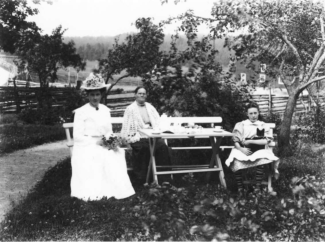 Calla Helena Sundbeck, Group picture with three women of different ages sitting in a garden furniture at Örserum’s school. On the left the teacher Hildevard Valler in bright dress and hat, next to her mother Sofia Johansson with crocheted lace over her shoulders and on the right Hildegard’s foster girl Tea Walén. The roa…